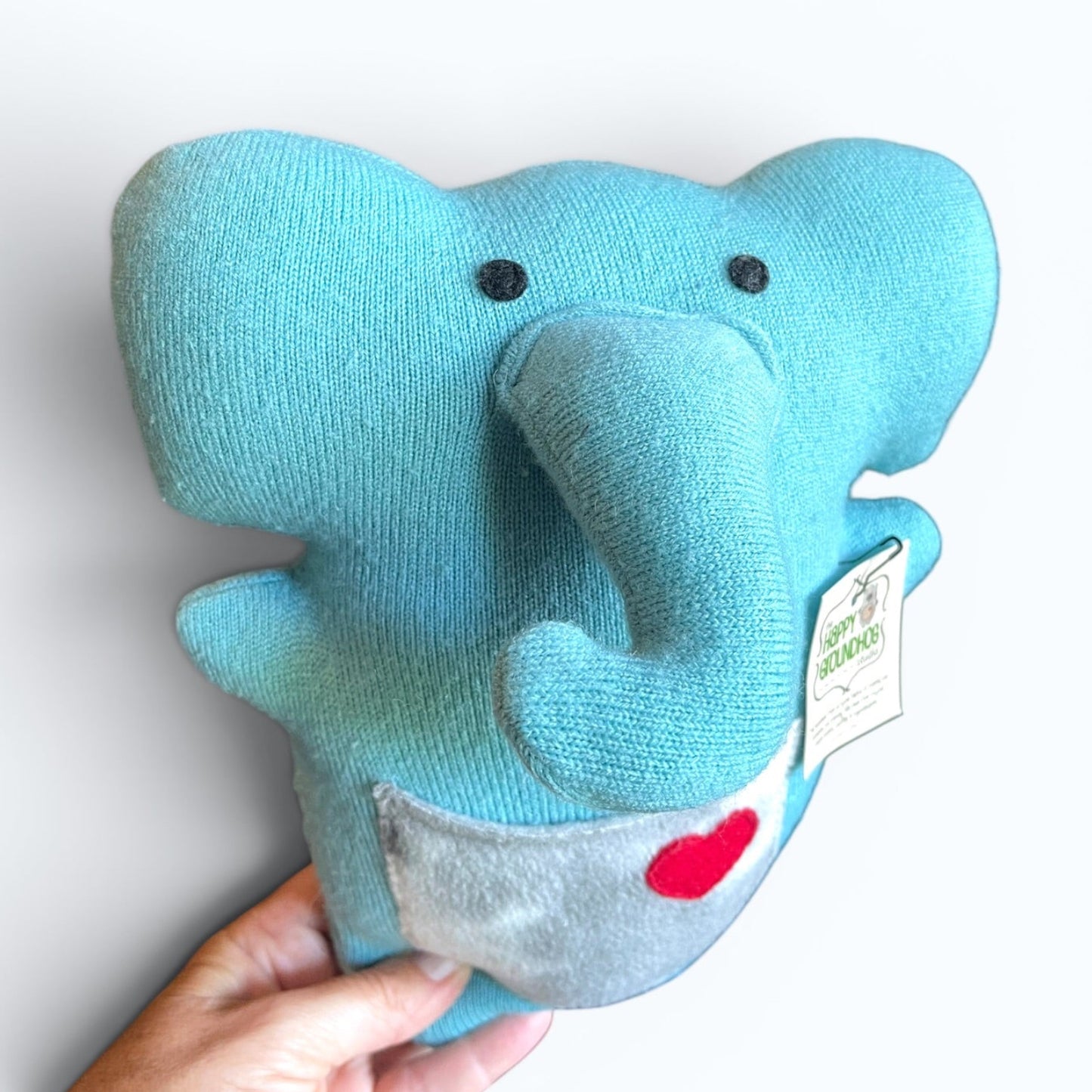 A light blue knit elephant plush toy being held by a hand against a plain white background. The toy features black felt  eyes, a small red heart on a gray pocket and a hanging tag The reads The Happy Groundhog Studio