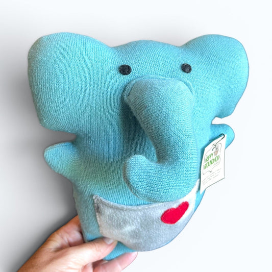 A light blue knit elephant plush toy being held by a hand against a plain white background. The toy features black felt  eyes, a small red heart on a gray pocket and a hanging tag The reads The Happy Groundhog Studio