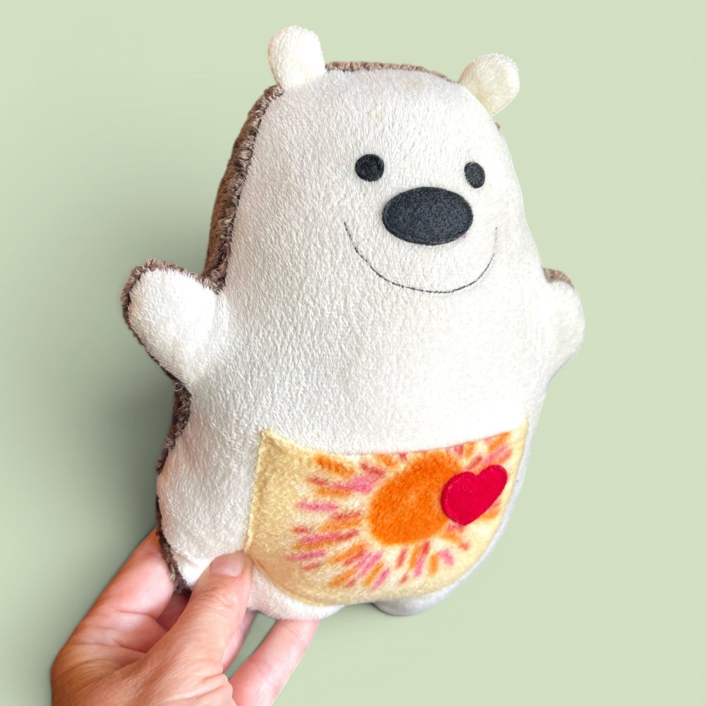 A hand holding a handmade plush hedgehog against a solid pale green background. The hedgehog is cream-colored with a brown fabric back,  black felt eyes and nose, and a yellow-orange sun and red heart sewn on its belly.