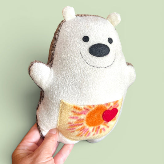 A hand holding a handmade plush hedgehog against a solid pale green background. The hedgehog is cream-colored with a brown fabric back,  black felt eyes and nose, and a yellow-orange sun and red heart sewn on its belly.