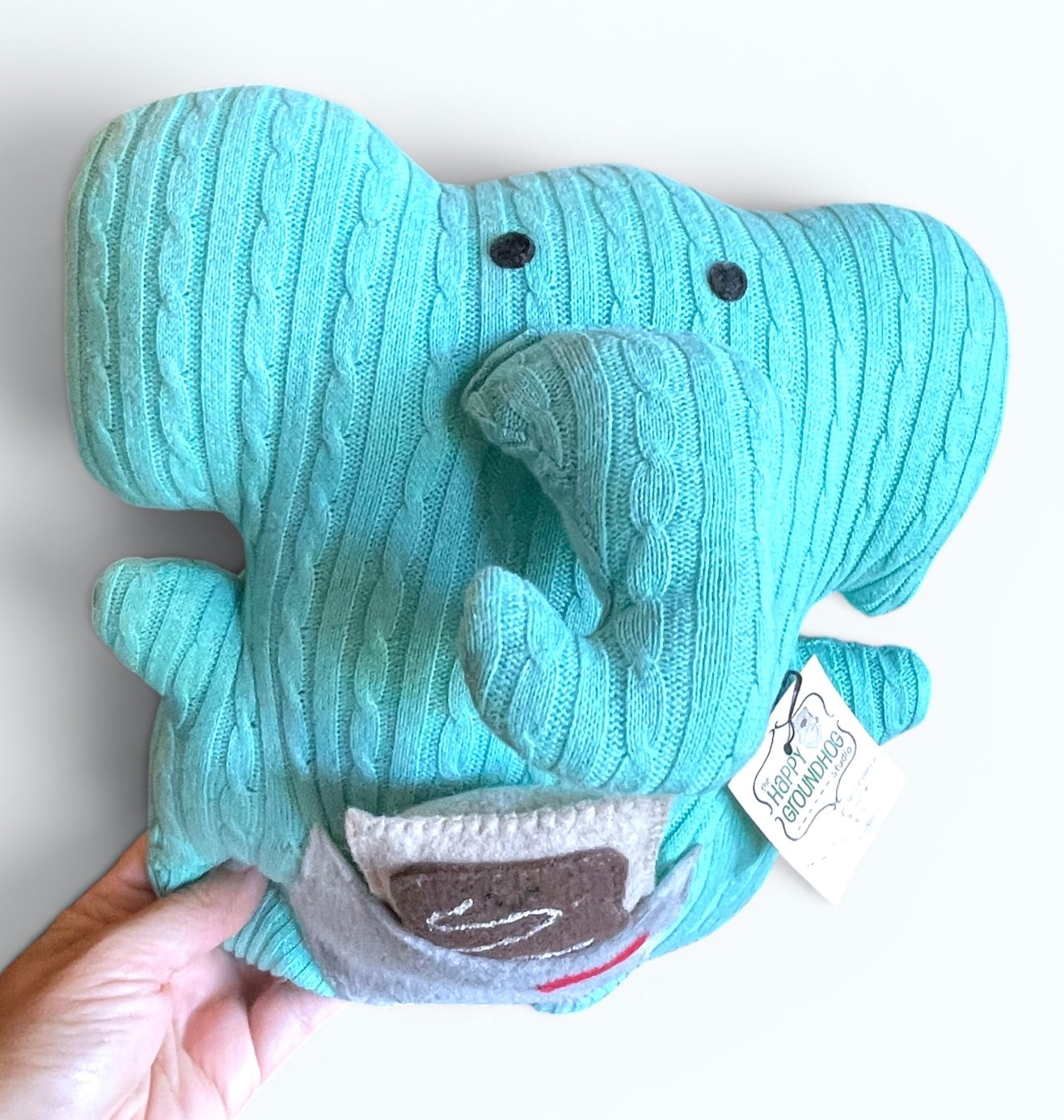 A light blue cable-knit elephant handmade plushie being held by a hand against a plain white background. The plushie features black felt  eyes, a gray fabric pouch, holding a felt poptart