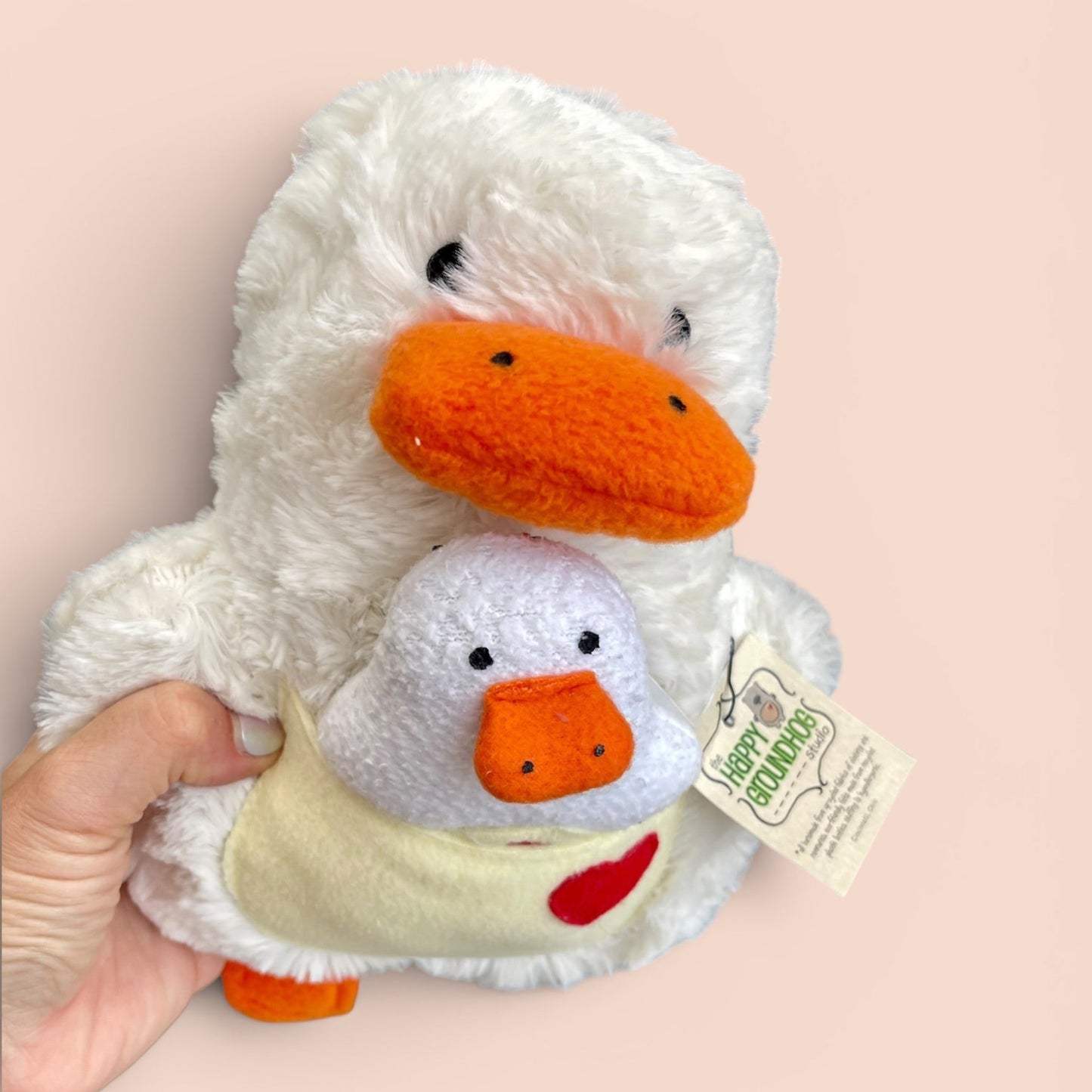 A hand holding a plush white goose toy with a large orange beak and a smaller plush duck nestled inside, against a solid pale peach background. A hangtag reading 'Happy Groundhog Studio' is attached to the side.