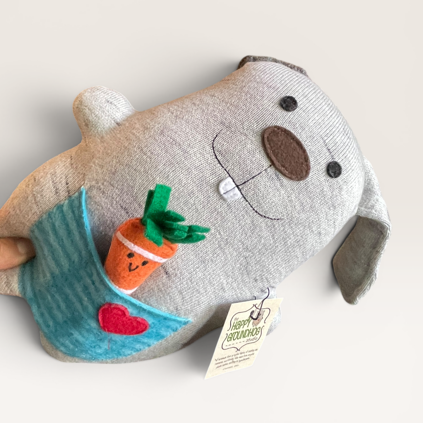 A plush bunny  in light heather gray up cycled fabric, featuring embroidered facial details, a blue striped pocket with a small orange carrot and red heart applique, and a hanging paper tag.