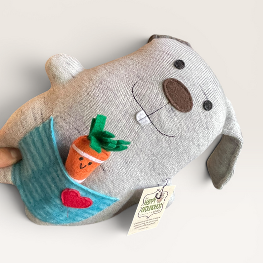 A plush bunny  in light heather gray up cycled fabric, featuring embroidered facial details, a blue striped pocket with a small orange carrot and red heart applique, and a hanging paper tag.