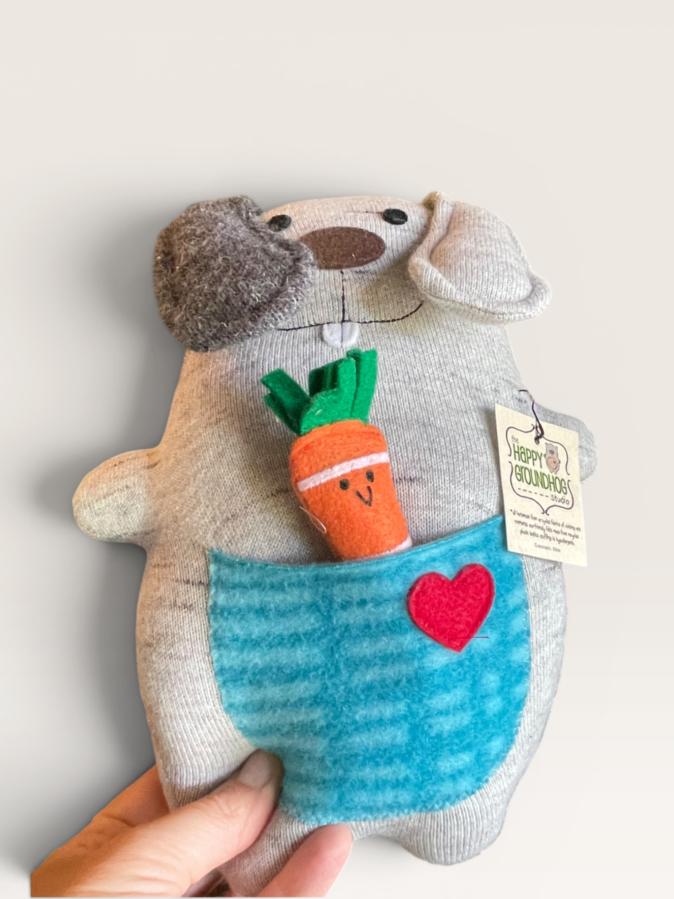 A plush stuffed animal toy shaped like a gray animal character with a blue striped front pocket, red heart detail, and a small carrot accessory, held by a hand against a plain light background.