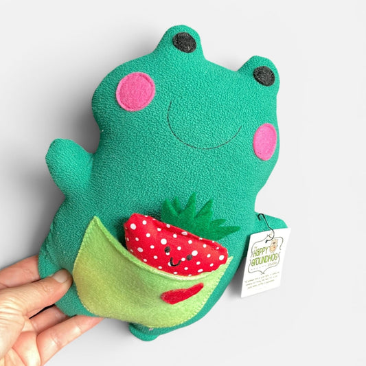 A green plush handmade frog toy held by a hand against a light gray background. The frog has pink circular cheeks, black felt eyes, a stitched smile, and a green pocket holding a red strawberry with white polka dots.