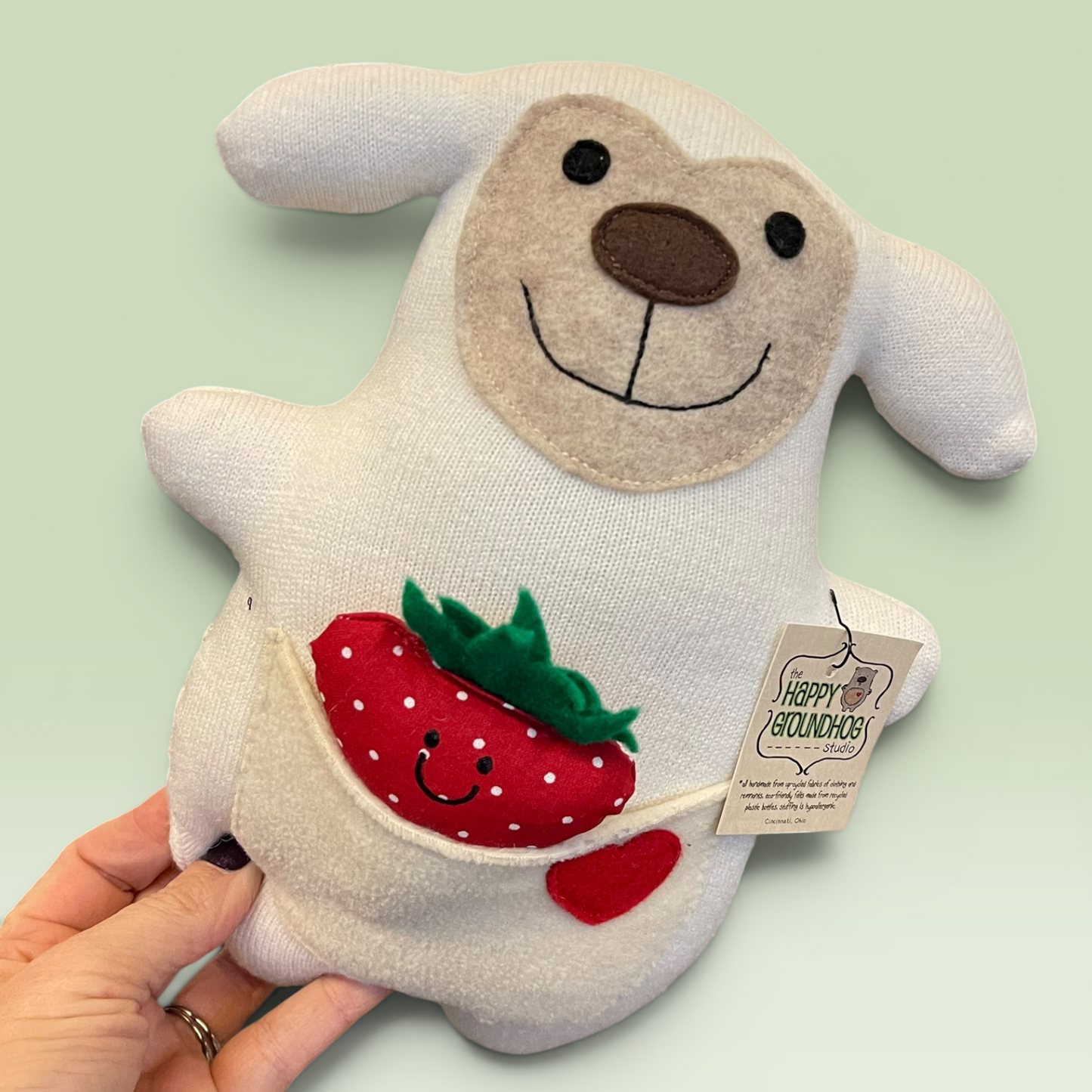 A hand holds a white sheep , a brown nose, embroidered smile, and a front pocket holding a red strawberry and a small red heart. A hangtag reads Happy Groundhog Studio