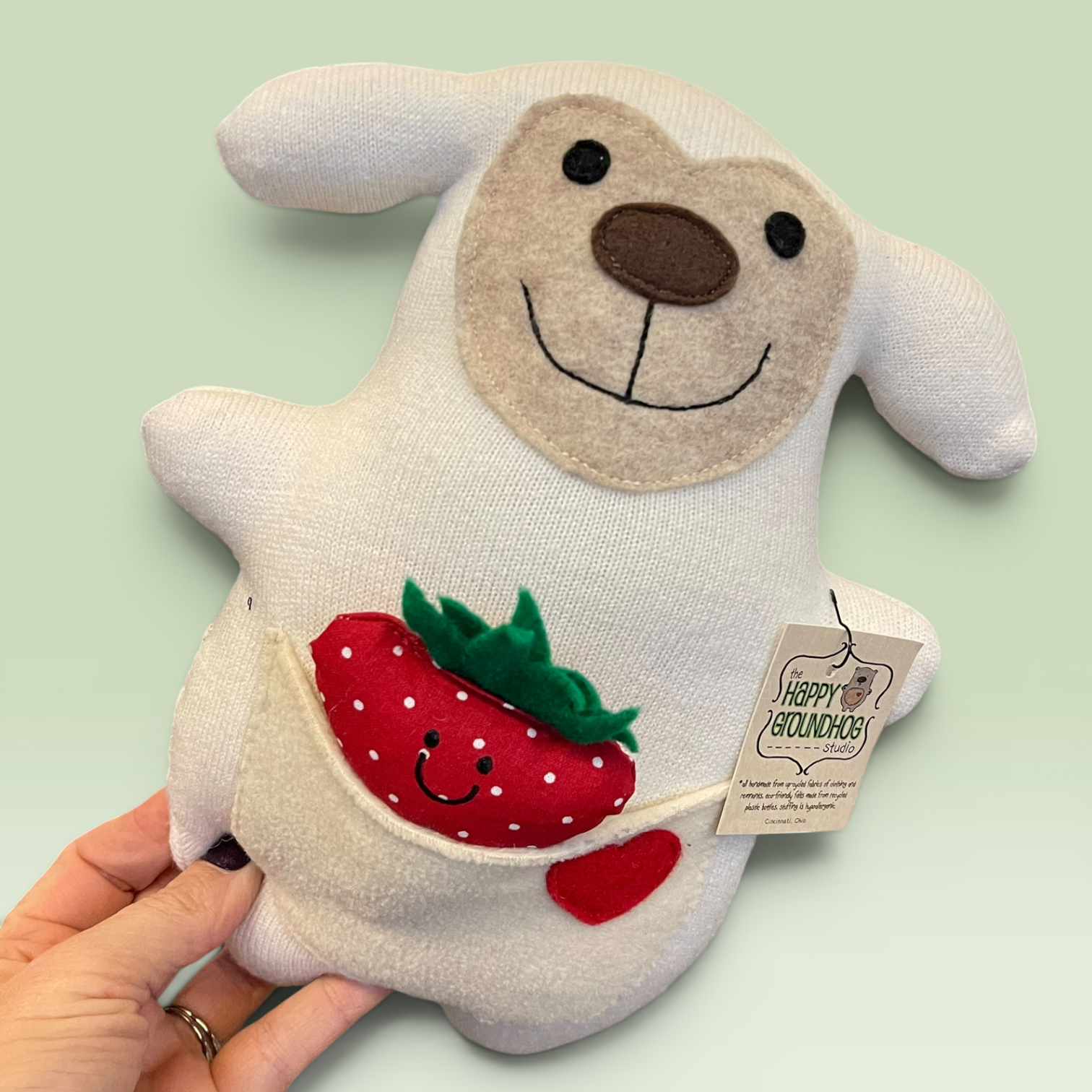 A hand holds a white sheep , a brown nose, embroidered smile, and a front pocket holding a red strawberry and a small red heart. A hangtag reads Happy Groundhog Studio
