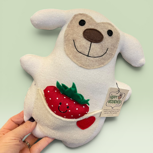 A hand holds a white sheep , a brown nose, embroidered smile, and a front pocket holding a red strawberry and a small red heart. A hangtag reads Happy Groundhog Studio