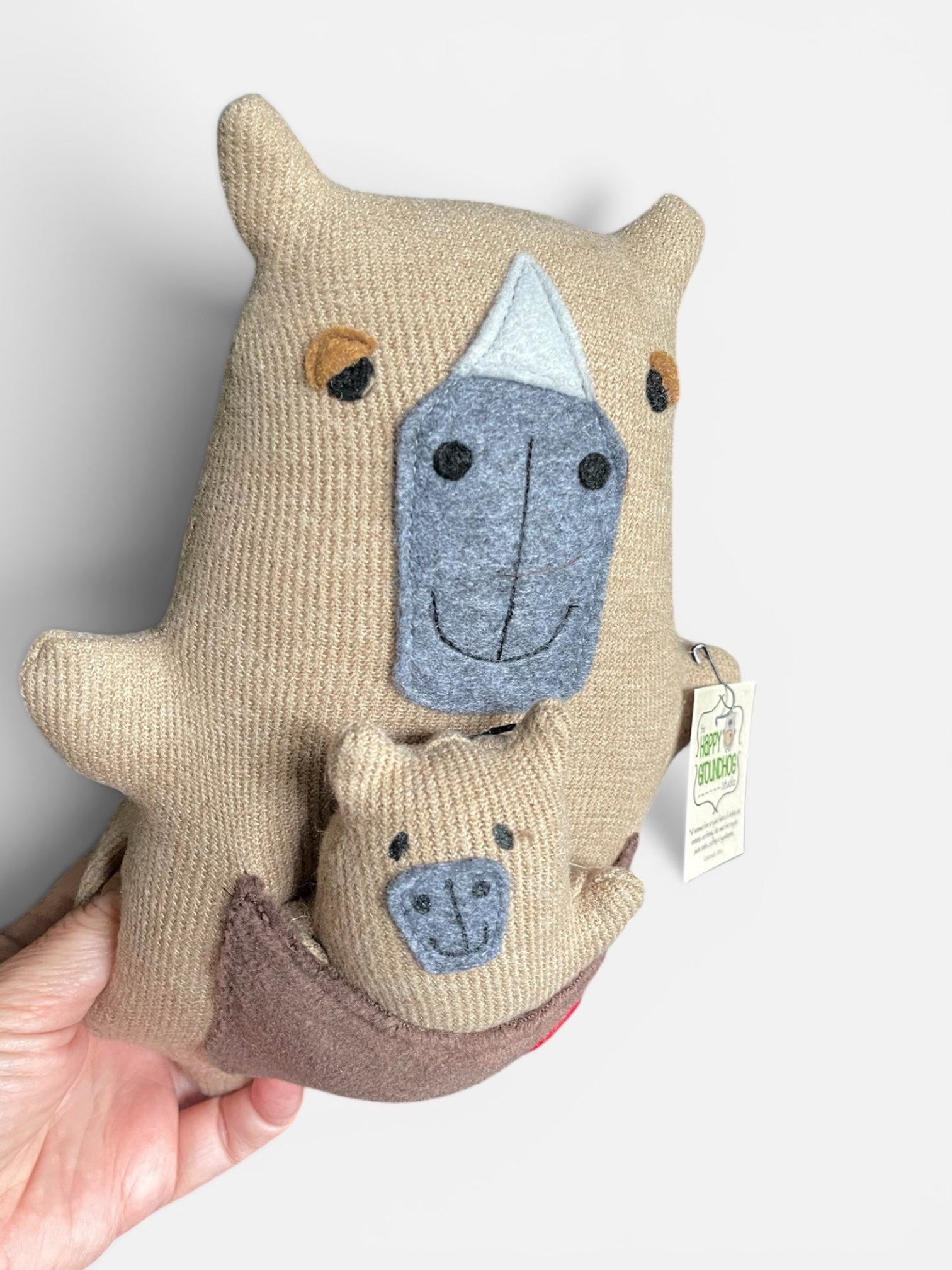 A hand holding a handmade capybara with a  smaller matching capybara nested inside its brown pouch, shown against a plain light background.